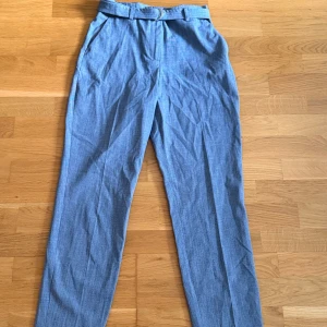 Grå kostymbyxor med bälte - Snygga grå kostymbyxor med diskret rutigt mönster och tillhörande bälte med metallspänne. Byxorna har hög midja, raka ben och dragkedja framtill. Perfekta för en clean och stilren look. De ser blåa ut med tanke på ljuset men har bifogat (bild 2) hur de egentligen ser ut. 