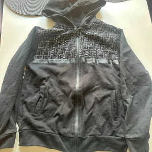 Svart hoodie med mönstrad mesh - Svart hoodie med huva och hel dragkedja framtill. Över bröstet finns ett coolt geometriskt mönster i mesh med vita detaljer som ger en unik look. Tröjan har två fickor på sidorna och är långärmad. Perfekt för dig som gillar streetwear och vill sticka ut. (Liten i storleken)