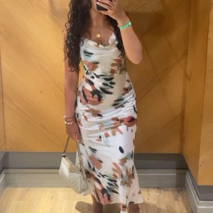 Vit långklänning med abstrakt mönster - Säljer en vit långklänning med snyggt abstrakt mönster i svart, beige och rostiga toner. Klänningen har smala axelband, cowlneck-ringning och en figurnära passform som ger en elegant siluett. Perfekt för dig som vill sticka ut med stil.