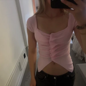 Ljusrosa Zara topp med knappar - Säljer denna trendiga slutsålda Zara blus som är ljusrosa , croppad topp med korta ärmar och snygg v-ringning. Toppen har dekorativa knappar framtill och ett draperat tyg som ger en cool twist. Perfekt för dig som gillar trendiga och feminina plagg.