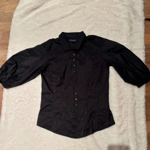 Svart randig skjorta The Shirt Factory - Svart skjorta från The Shirt Factory med tunna vita ränder. Skjortan har trekvartsärmar med lätt puff och knäppning framtill. Snygg krage och figurnära passform. Tillverkad i 100% polyester, perfekt för en stilren look.