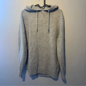 Grå merinoull hoodie - Mysig grå stickad hoodie med huva och snörning framtill. Tröjan har lång ärm och ribbade muddar vid ärmslut och nederkant. Perfekt för lager-på-lager och chill dagar. Använd fåtal gånger och i stort sett i nytt skick! Bara att höra av sig vid frågor och funderingar!