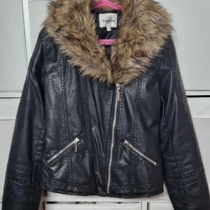 Svart skinnjacka med fuskpäls KappAhl - Svart skinnjacka från KappAhl med snyggt fuskpälsfoder i kragen. Jackan har dragkedja framtill, två snedställda fickor med dragkedjor och cool bikerstil. Perfekt för dig som vill ha en edgy look med extra detaljer. Storlek 152 men passar xs/s