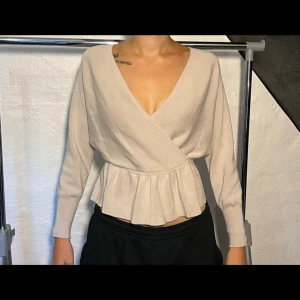 Omlottopp med öppen rygg från Bubbleroom  - Superfin beige omlottopp från Bubbleroom i mjukt stickat material. Toppen har v-ringning, lång ärm och volangdetalj i midjan. Snygg öppen rygg med band upptill som håller axlarna på plats. 