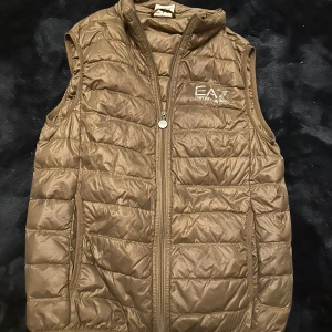 Militär grön dunväst från Emporio Armani - Snygg militär grön dunväst från Emporio Armani med quiltad design och dragkedja framtill. Västen har hög krage och logga på bröstet. Perfekt för lager-på-lager och ger en clean look till din outfit.