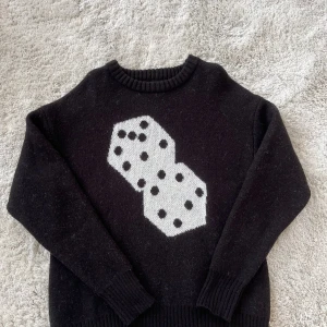 Svart stickad Stüssy tröja med tärningsmotiv - Svart stickad tröja från Stüssy i storlek L med ett stort vitt tärningsmotiv på framsidan. Tröjan har rund hals, ribbade muddar och är långärmad. Materialet är mjukt och värmande, perfekt för kyliga dagar. Snygg streetwear-vibe och unik design.
