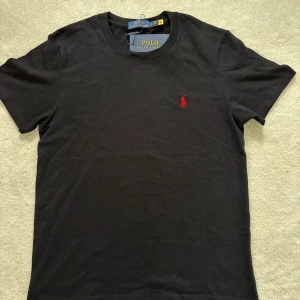 Svart t-shirt från Polo Ralph Lauren - Svart t-shirt från Polo Ralph Lauren med klassisk rund hals och korta ärmar. På bröstet finns det ikoniska röda broderade Polo-logotypen. Tillverkad i mjuk bomull för skön komfort och stilren look.