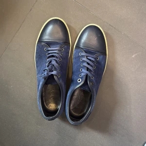 Blå mocka sneakers från Lanvin - Snygga sneakers från Lanvin i mörkblå mocka med rund tå och snörning. Skorna har en vit platt sula och detaljer i svart skinn på tån och hälen. Perfekt för dig som gillar stilrena och exklusiva sneakers med lyxig känsla.