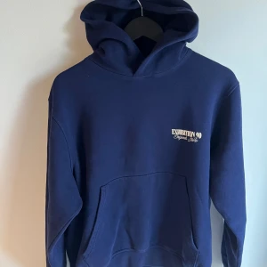 Mörkblå hoodie Exhibition 90 - Snygg mörkblå hoodie från Exhibition 90 med vit textprint både fram och bak. Klassisk känguruficka och stor huva. Perfekt för dig som gillar streetwear och vill sticka ut med coola tryck. Mjuk och skön bomullskänsla.
