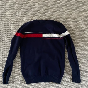 Stickar Mörkblå Tommy Hillfigger tröja - Snygg mörkblå stickad tröja med långa ärmar och coola röda och vita linjer över bröstet. Tröjan har rund halsringning och ribbade muddar vid ärmslut och nederkant. Perfekt för dig som gillar stilrena men ändå unika plagg.