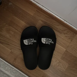 Svarta sandaler från The North Face - Svarta sandaler från The North Face med vit logga på ovansidan. Modellen har en bred rem över foten och platt sula med mönstrad undersida för extra grepp. Tillverkade i syntetmaterial, perfekt för chill dagar och sommarhäng.