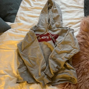 Grå Levi's hoodie med röd logga - Grå hoodie från Levi's med stor röd logga på bröstet. Tröjan har huva med snörning, magficka och långa ärmar. Mjuk och skön bomullsmix, perfekt för chill dagar. Passformen är normal och modellen är klassisk. Pris går att diskuteras💞