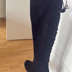 Svarta mockastövlar med snörning, strl 39 - Snygga svarta stövlar i mocka med snörning längs sidan och rund tå. Klacken är cirka 6 cm hög och ger en cool siluett. Stövlarna går upp över knäet och har en mjuk, lyxig känsla. Perfekt för dig som gillar statement boots med lite edge.