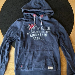 Blå hoodie från NZA Auckland  - Säljer en mörkblå hoodie från NZA Auckland i storlek XS. Tröjan har tryck och broderad detalj framtill, snörning i huvan med röda detaljer och en stor magficka. Materialet är mjuk bomull och polyester, perfekt för chill dagar. Snygg och bekväm passform.