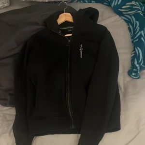 Svart hoodie från Sail Racing - Svart hoodie från Sail Racing med huva och dragkedja framtill. Tröjan har en diskret logga på bröstet och en praktisk ficka med dragkedja. Mjuk och skön, perfekt för chill dagar. Långärmad och med ribbade muddar för extra komfort. Jag har andvönt den max 10 gånger den har inga skador 