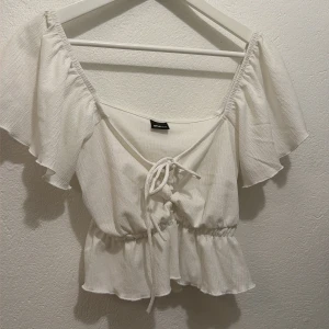 Vit blus med volang  - Supersöt vit blus från Gina Tricot med volangkant och korta, fladdriga ärmar. Toppen har snörning framtill och en croppad passform med resår i midjan för en snygg siluett. 🩷