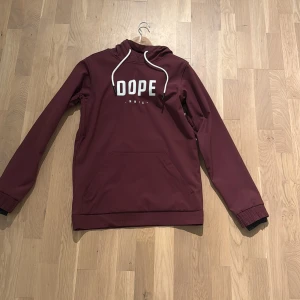 Vinröd skidjacka från Dope - Säljer en vinröd skidjacka från Dope med vit logga på bröstet och vita snören i huvan. Jackan har känguruficka framtill, långa ärmar och ribbade muddar. Perfekt för dig som gillar streetwear och vill ha en snygg och bekväm look i backen.