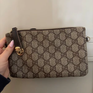 brun kuvertväska - Snygg kuvertväska i brunt med klassiskt GG-monogram över hela väskan. Väskan har guldfärgade detaljer, dragkedja upptill och en extra ficka med dragkedja på baksidan. Tillverkad i canvas med detaljer i skinn.