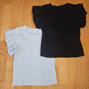 Två volangtopp från Gina Tricot - Säljer två snygga toppar från Gina Tricot i storlek 146/152. En är svart och en är ljusblå, båda med korta volangärmar och ribbad struktur. Perfekta för dig som gillar en söt men stilren look. 