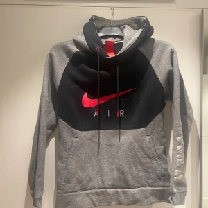 Nike Air hoodie i grått och svart - Nike Air hoodie i grått och svart med stor röd swoosh-logga på bröstet och tryck på ena ärmen. Tröjan har huva med snörning, känguruficka och är tillverkad i mjuk bomullsblandning. Perfekt för en sportig och chill stil.