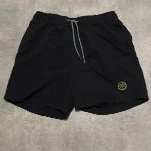 Svarta badshorts Jack & Jones S - Svarta badshorts från Jack & Jones med elastisk midja och grå snörning. På vänster ben finns en gul palm-logga. Shortsen är tillverkade i snabbtorkande syntetmaterial och har en klassisk, avslappnad passform. Perfekta för stranden eller poolen.