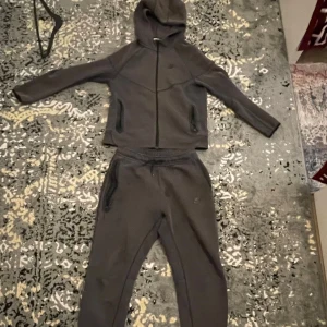 Mörkgrå Nike hoodie med dragkedja - Säljer en mörkgrå hoodie från Nike med hel dragkedja framtill och huva. Tröjan har långa ärmar, två fickor med dragkedja och en sportig look. Perfekt för chill eller träning. Matchande byxor medföljer, båda i mjukt material.använd över 5 gånger och under 20 