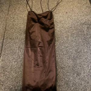 Brun långklänning med tunna axelband - Säljer en elegant brun långklänning med tunna axelband och rak passform. Klänningen har en slät, glansig yta som ger en lyxig känsla och är perfekt för dig som gillar minimalistisk stil.