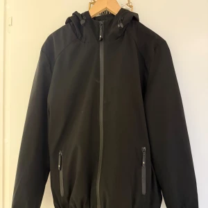 Svart vindjacka med huva - Svart vindjacka med huva och hel dragkedja framtill. Jackan har två fickor med dragkedja och elastisk nederkant. Enkel och stilren design som passar perfekt till vardags. Materialet känns lätt och smidigt. Pris kan diskuteras.