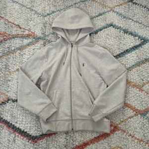Beige hoodie från Polo Ralph Lauren - Säljer en stilren ljusgrå hoodie från Polo Ralph Lauren i storlek S. Tröjan har luva med snörning, dragkedja framtill och två fickor. Klassisk logga broderad på bröstet. Perfekt för chilla dagar och enkel att matcha med jeans. Perfekt för dig som vill ha en stilren look