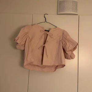 Beige blus med puffärm och knyt - Säljer en rosa blus med korta puffärmar och två söta knyt framtill. Blusen har en croppad passform och är tillverkad i ett lätt bomullsmaterial. Perfekt för dig som gillar voluminösa ärmar och romantiska detaljer.