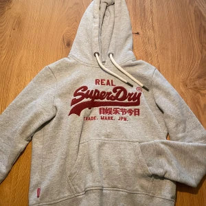 Grå Superdry hoodie med röd logga - Säljer en grå hoodie från Superdry med stor röd logotyp och japanska tecken på bröstet. Tröjan har huva med vita snören, känguruficka och ribbade muddar vid ärmar och nederkant. Perfekt för dig som vill ha en snygg, bekväm look.