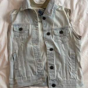 Ljusblå jeansväst med knappar - Snygg ljusblå jeansväst med klassisk krage och dekorativa svarta knappar framtill. Västen har två bröstfickor med lock och två sidofickor. Perfekt att slänga över en hoodie eller t-shirt, köpt i Polen så okänt märke! 🩷