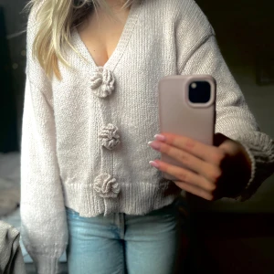 Beige stickad kofta med blommiga knappar - Superfin beige stickad kofta med v-ringning och stora blommiga knappar framtill. Koftan har lång ärm och en lite croppad passform som ger en trendig look. Perfekt att slänga över ett linne eller t-shirt för en mysig vibe.