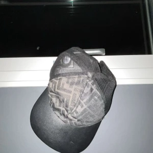 Svart meshkeps från Fendi - Snygg svart keps från Fendi med klassiskt FF-mönster framtill och mesh på sidorna och bak. Justerbar rem baktill med gul Fendi-logga. Perfekt accessoar för en streetwear-look. Materialet är polyester och mesh.