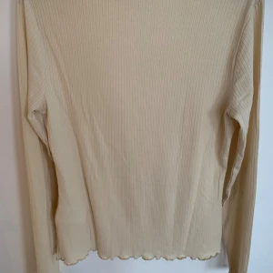 Beige ribbad långärmad topp - Säljer en beige långärmad topp med ribbad struktur och lätt vågad kant vid hals och nederdel. Toppen har en enkel och clean look som är lätt att matcha med olika outfits. Perfekt för dig som gillar stilrena basplagg.