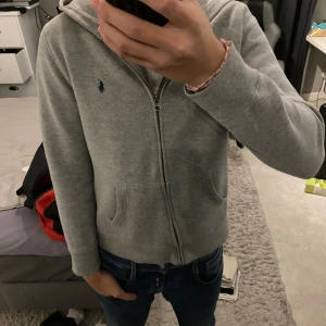 Grå hoodie från Polo Ralph Lauren - Säljer en stilren grå hoodie från Polo Ralph Lauren med dragkedja och huva. Tröjan har två fickor framtill och klassisk blå logga broderad på bröstet. Perfekt för chill dagar och enkel att matcha med jeans eller joggers.