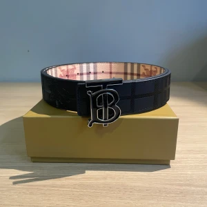 Svart Burberry bälte med rutig insida - Stilrent svart bälte från Burberry med klassiskt rutigt mönster på insidan och en stor metallspänne med Burberrys ikoniska monogram. Bältet är tillverkat i skinn och levereras i originalförpackning med dustbag och certifikat.