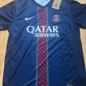 PSG hemmatröja 25/26 matchtröja Nike marinblå - Paris Saint-Germain fotbollströja från Nike i marinblått med rött och vitt geometriskt mönster längs mitten. PSG-logga på bröstet och Qatar Airways tryck. Kortärmad och tillverkad i lätt polyester som andas. Perfekt för dig som vill representera PSG på planen eller läktaren.