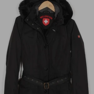 Svart parkajacka från Wellensteyn - Svart parkajacka från Wellensteyn med avtagbar huva och fuskpäls, quiltat innerfoder i brons, bälte i midjan och dragkedja samt knappar framtill. Jackan har flera fickor och snygga detaljer som ger en clean och stilren look.