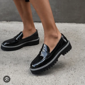 Jo Mercer Australia Svarta chunky loafers i lack - Säljer ett par svarta loafers med chunky platt sula och grovt mönstrad yttersula. Skorna är i glansigt lackmaterial med dekorativ söm runt sulan och klassisk pennyloafers-detalj framtill. Perfekt för dig som gillar att sticka ut med din stil.