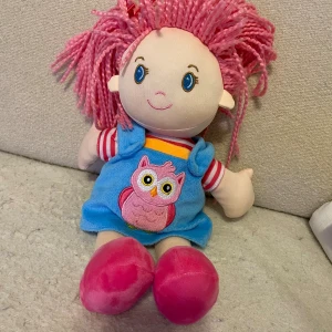 Mjukdocka med rosa hår -  Mjukdocka med rosa hårSöt mjukdocka med rosa garnhår och en blå klänning med en uggla på. Dockans längd är cirka 32cm