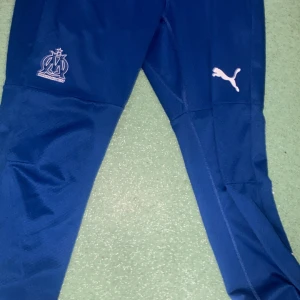 Blå träningsbyxor Puma Marseille - Snygga blå träningsbyxor från Puma med Olympique de Marseille-logga på ena benet och Puma-logga på det andra. Byxorna har smal passform, ventilerande meshdetaljer och är gjorda i polyester. Perfekta för fotbollsträning eller chill.