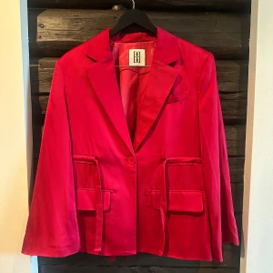 Röd kostym med kavaj och utställda byxor - Säljer min fina röda kostym från Marlene Birger - aldrig snväbd. Kavajen har knappar, fickor med lock och dekorativa banddetaljer i ryggen. Byxorna har normalhög midja och vida ben. Materialet är lyxigt i sidenkvalitet (acetat). Nypris 4600 sek. 