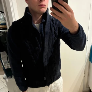 Mörkblå stickad väst Tommy Hilfiger - Säljer en mörkblå stickad väst från Tommy Hilfiger i storlek S. Västen har grov kabelstickning framtill, hög ribbad krage och knäppning med knappar. Insidan har rutigt foder i rött och grönt. Perfekt för lager-på-lager och snygg till jeans. Storlek S, men sitter som xs. Lätt urtvättad men bra skick 