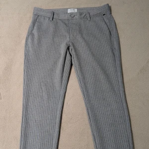 Grå randiga kostymbyxor Only & Sons - Snygga grå kostymbyxor från Only & Sons med tunna vita ränder. Byxorna har normal passform, bälteshällor, framfickor och bakfickor med knapp. Perfekta för en clean och stilren look. Materialet känns mjukt och lätt, vilket gör dem bekväma hela dagen för jobbet eller för speciella tillfällen