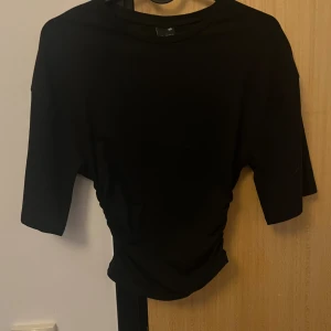 Svart topp med tajt midja  - Svart topp med halvlånga ärmar och rynkade sidor som ger en snygg siluett. Enkel och stilren design, perfekt att matcha med jeans eller kjol. Mjuk och stretchig känsla.