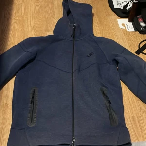 Mörkblå Nike hoodie med dragkedja - Snygg mörkblå hoodie från Nike med hel dragkedja och två fickor med dragkedja framtill. Tröjan har huva och diskret Nike-logga på bröstet. Perfekt för dig som gillar sportig och clean stil.