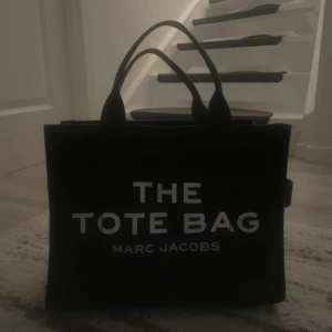 Svart tote bag från Marc Jacobs - Snygg svart tote bag från Marc Jacobs med tydlig logga framtill och cool patch på baksidan. Väskan är fyrkantig med dubbla handtag och har en praktisk innerficka med dragkedja. Perfekt storlek för att få plats med allt du behöver.
