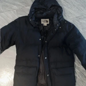 Svart dunjacka från The North Face - Säljer en svart dunjacka från The North Face med huva och stora fickor framtill. Jackan har dragkedja och tryckknappar, perfekt för kalla dagar. Jackan är i bra skick, säljer den billigt då det är lite smink på kragen som syns på bilderna, detta bör gå bort med våtservett eller kemtvätt ❤️