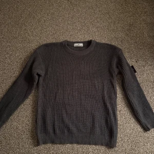 Stone Island tröja - Säljer en mörkgrå stickad tröja från Stone Island med klassisk rund hals och ribbade muddar. Tröjan har lång ärm och den ikoniska Stone Island patchen på vänster ärm. Perfekt för dig som gillar streetwear och snygga detaljer.